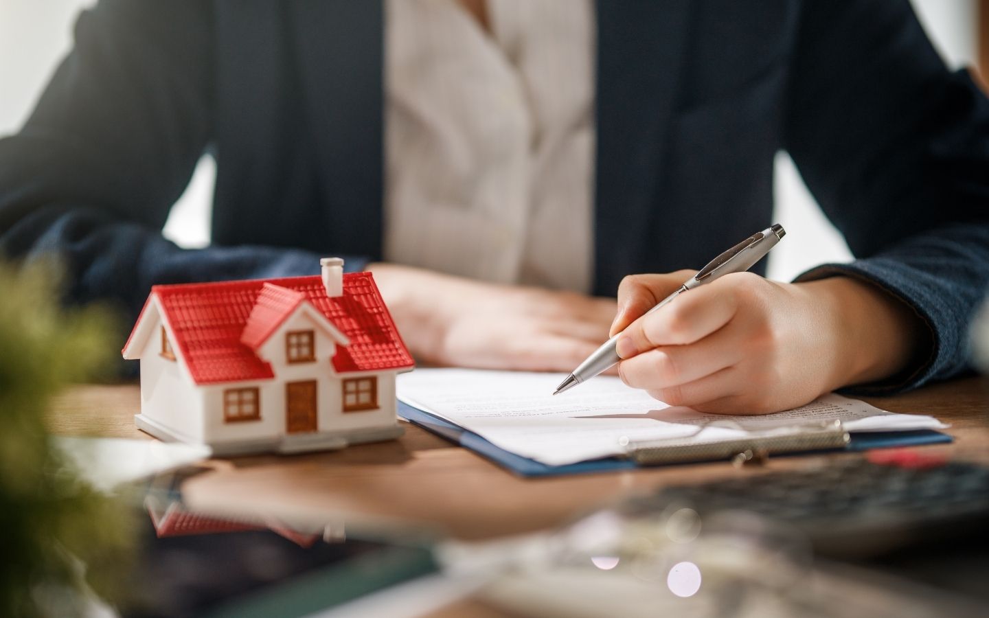 A person in a suit holding a silver pen to sign a contract on a clipboard next to a miniature house model on a wooden desk.