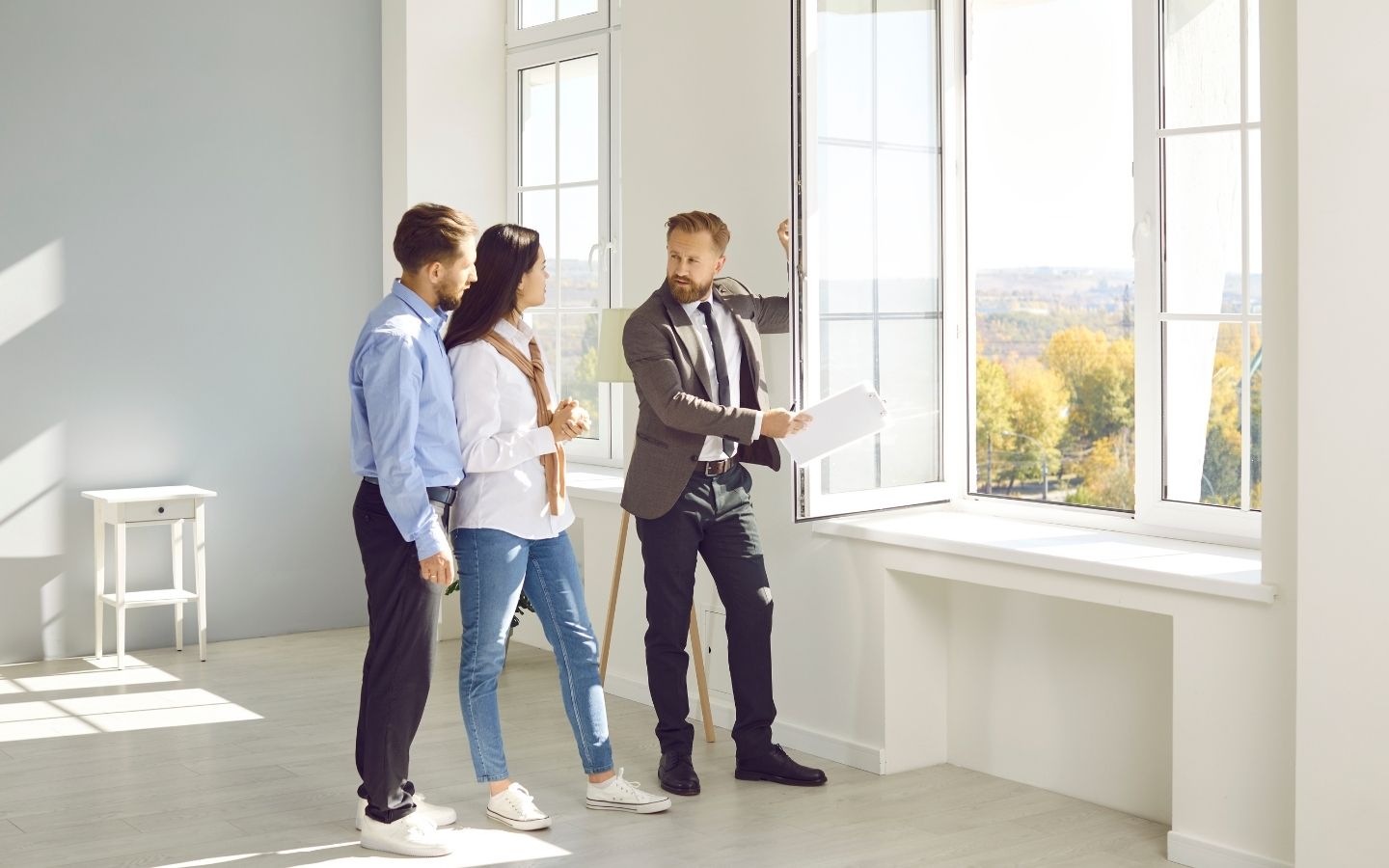 A real estate agent in a blazer holding documents on a clipboard while showing a bright, empty apartment room to a young couple near a large window.