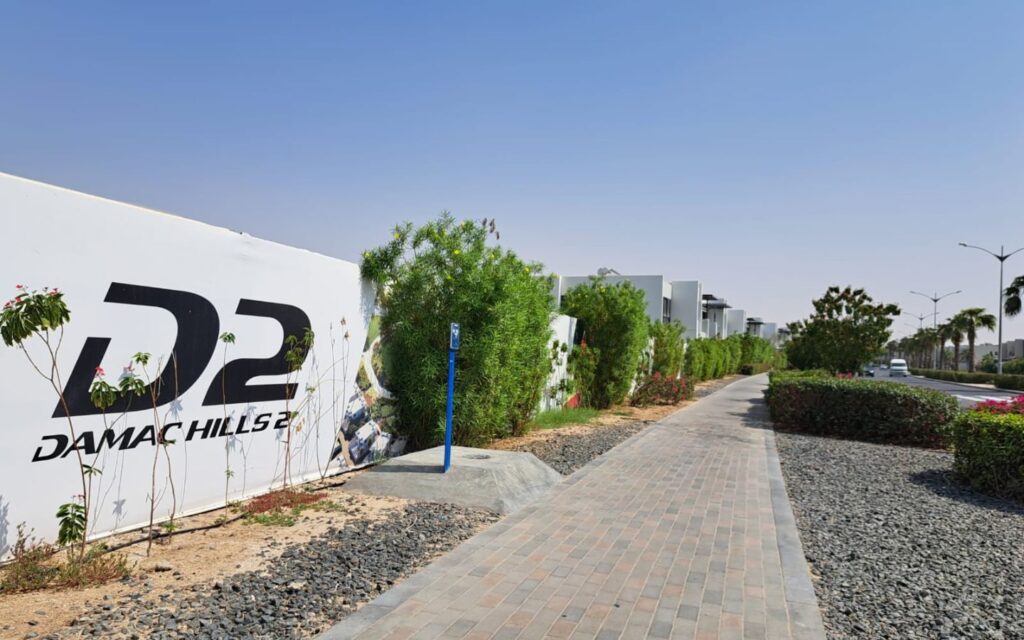 White wall displaying the D2 DAMAC HILLS 2 logo along a paved pedestrian walkway lined with green bushes