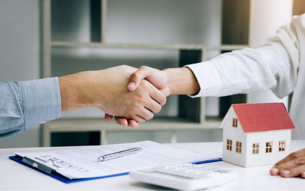 Real estate agents shaking hands over property contract with house model and documents on desk