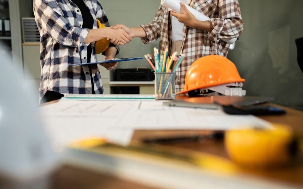Professionals shaking hands during a building project discussion with safety helmets and plans on a desk.
