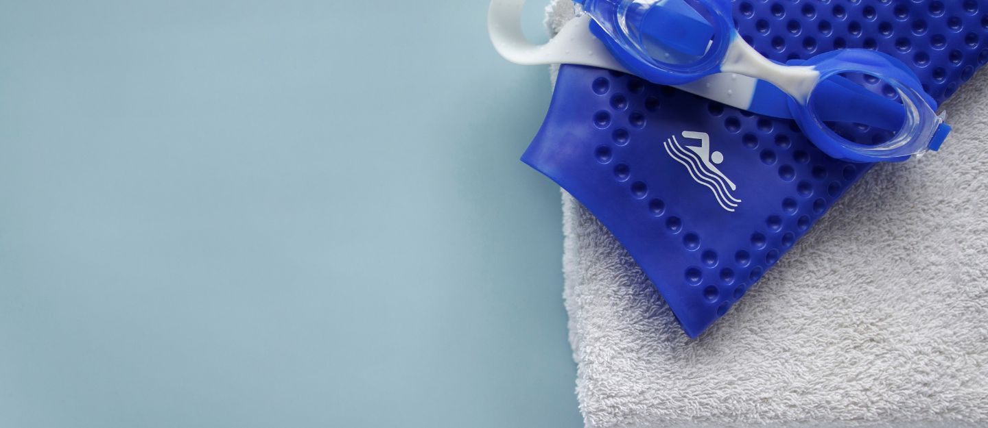 Blue swimming goggles and a matching blue silicone swim cap resting on top of a folded white towel
